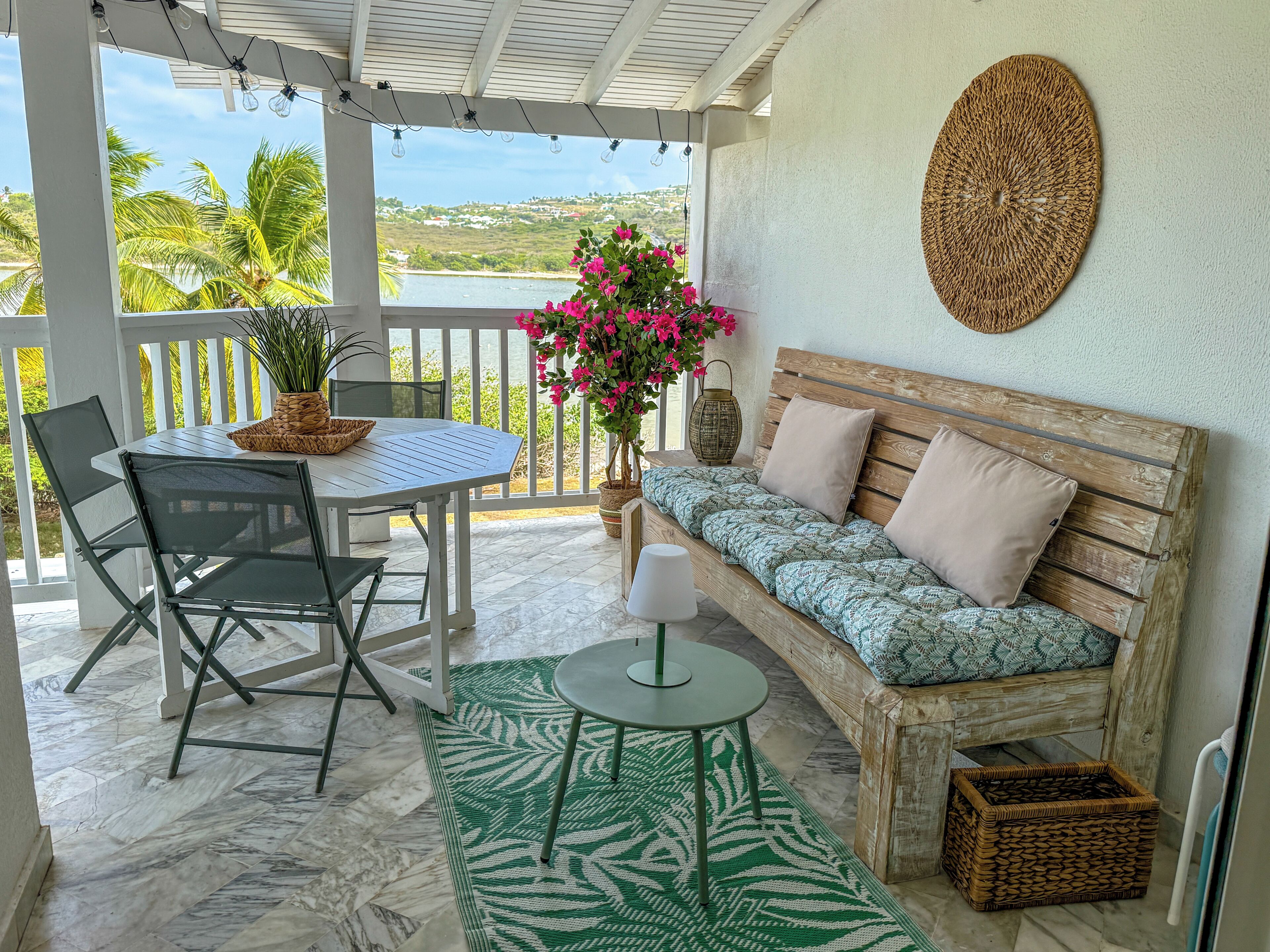 A covered outdoor seating area with a table, chairs, a sofa, a potted plant, and a view of the landscape.
