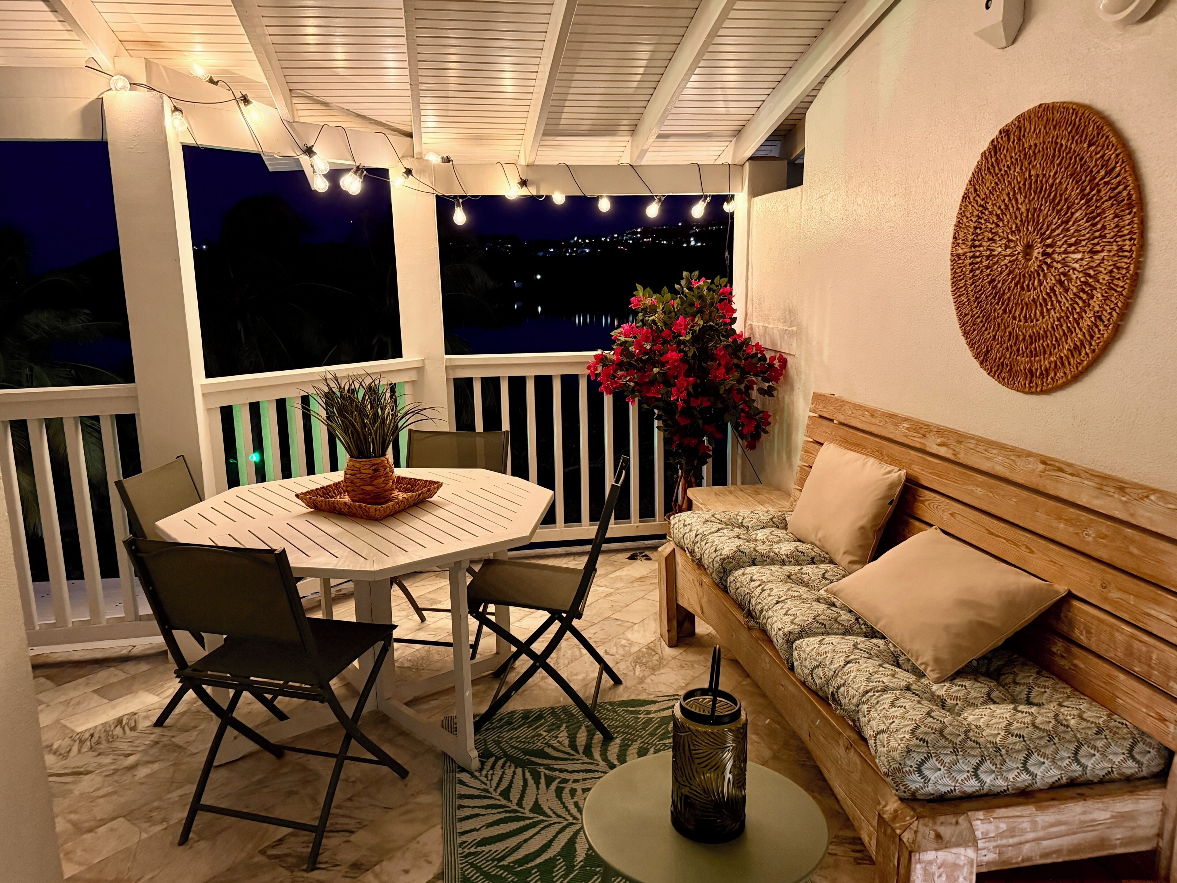 Un balcon avec une table blanche, des chaises, un canapé en bois, un élément décoratif mural et une plante en pot.