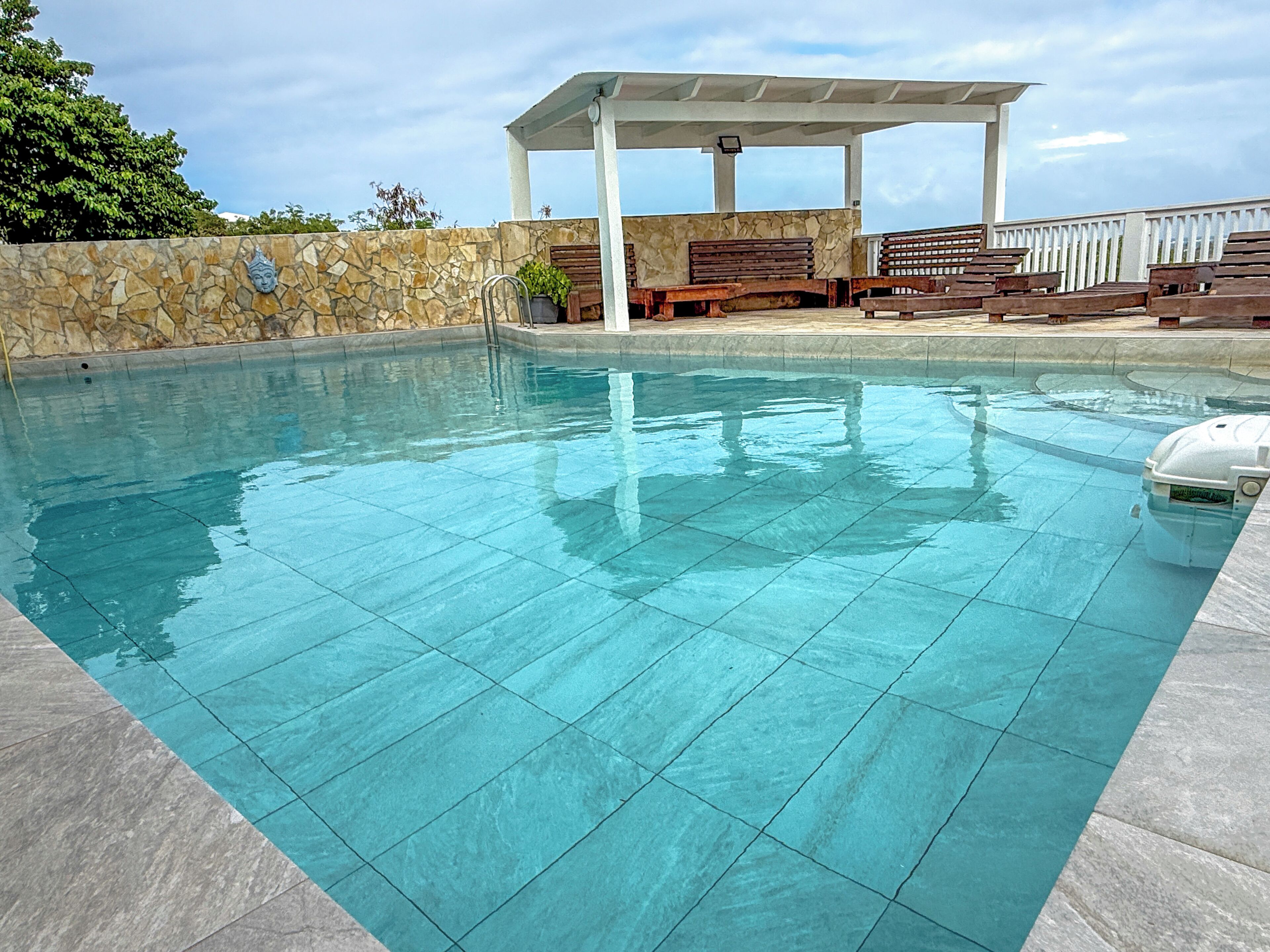 A swimming pool with a stone wall, a wooden deck, and a covered seating area.