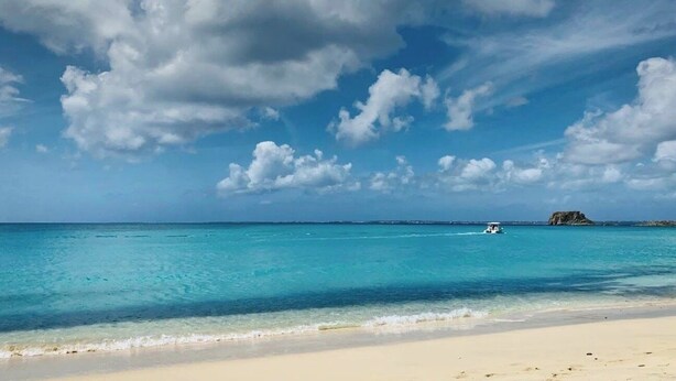 Een strand met helder turquoise water, een boot en een eiland in de verte.