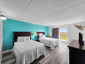 A hotel room with two beds, a desk, a lamp, and a view of the outdoors.