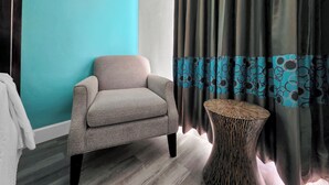 Een kamer met een blauwgroene wand, een grijze fauteuil, een gordijn met een patroon en een ronde houten tafel.