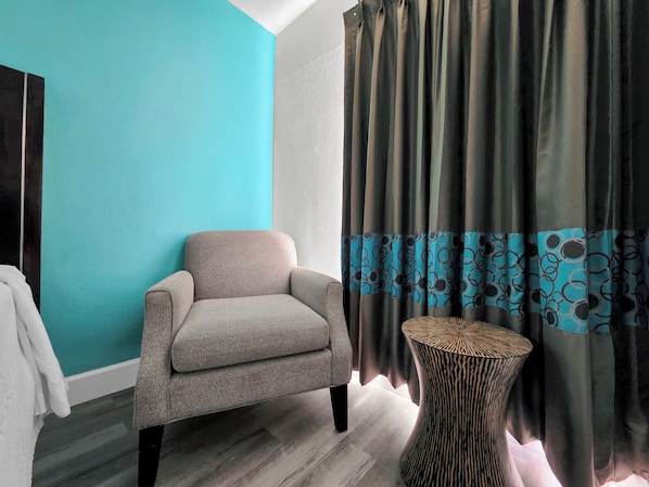 A room with a teal wall, a grey armchair, a patterned curtain, and a round wooden table.