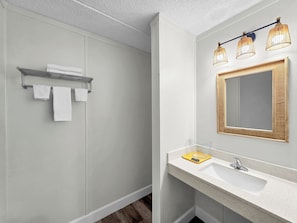 Un baño moderno con ducha, un lavamanos blanco con espejo de marco dorado y un perchero con tres toallas.