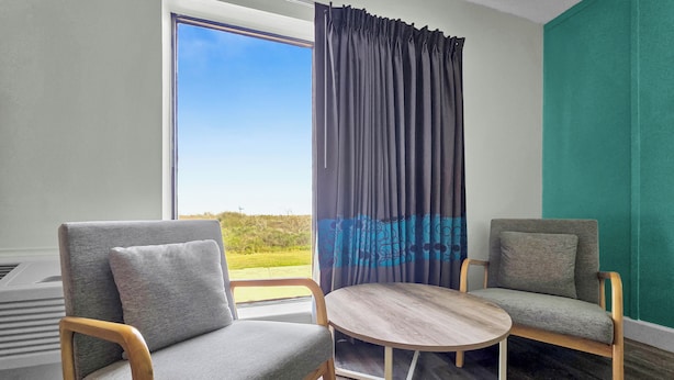 Une pièce comprenant deux fauteuils gris à revêtement en tissu, une table ronde en bois et une vue sur l’extérieur grâce à une grande fenêtre ornée de rideaux foncés.