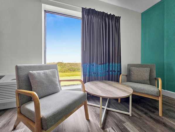 A room with two grey upholstered chairs, a round wooden table, and a view of the outdoors through a large window with dark curtains.
