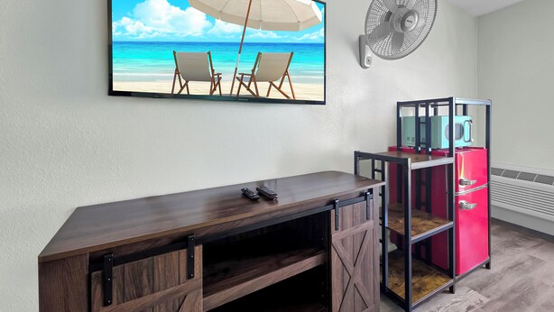 Ein Zimmer mit einem Strandgemälde, einem Ventilator, einem roten Kühlschrank und einem Holztisch.