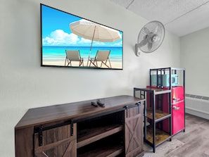 A room with a beach-themed painting, a fan, a red refrigerator, and a wooden desk.