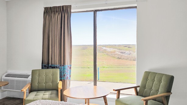 A hotel room with a bed, two armchairs, a small round table, a window with a view of a grassy field, and an air conditioning unit.