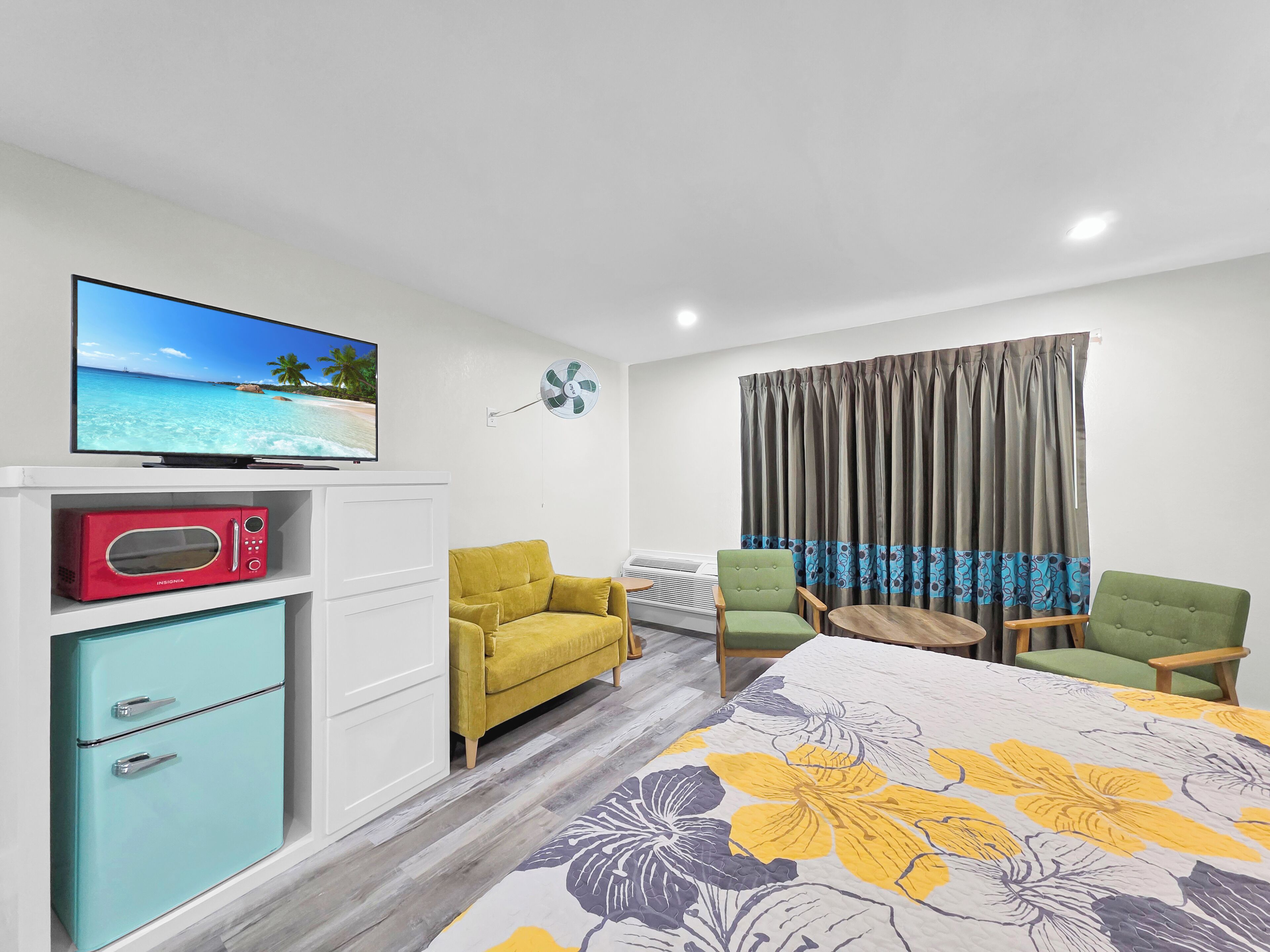 A hotel room with a television, a microwave, a sofa, a chair, a bed with a floral bedspread, and a window with curtains.