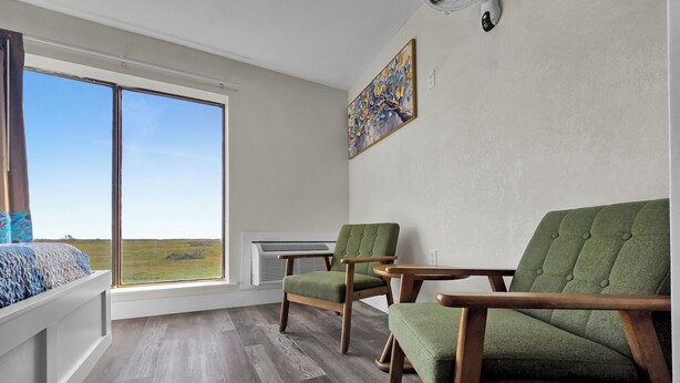 A room with a large window, two green upholstered chairs, a wooden table, and a painting on the wall.