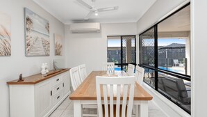 Un comedor con mesa de madera y sillas blancas, un equipo de aire acondicionado y vista a una piscina a través de un amplio ventanal.