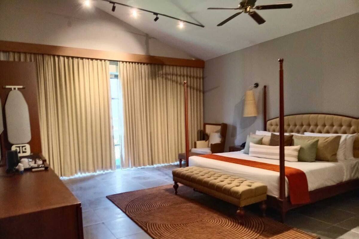 A bedroom with a four-poster bed, a desk, a chair, a bedside table, a lamp, and a ceiling fan.