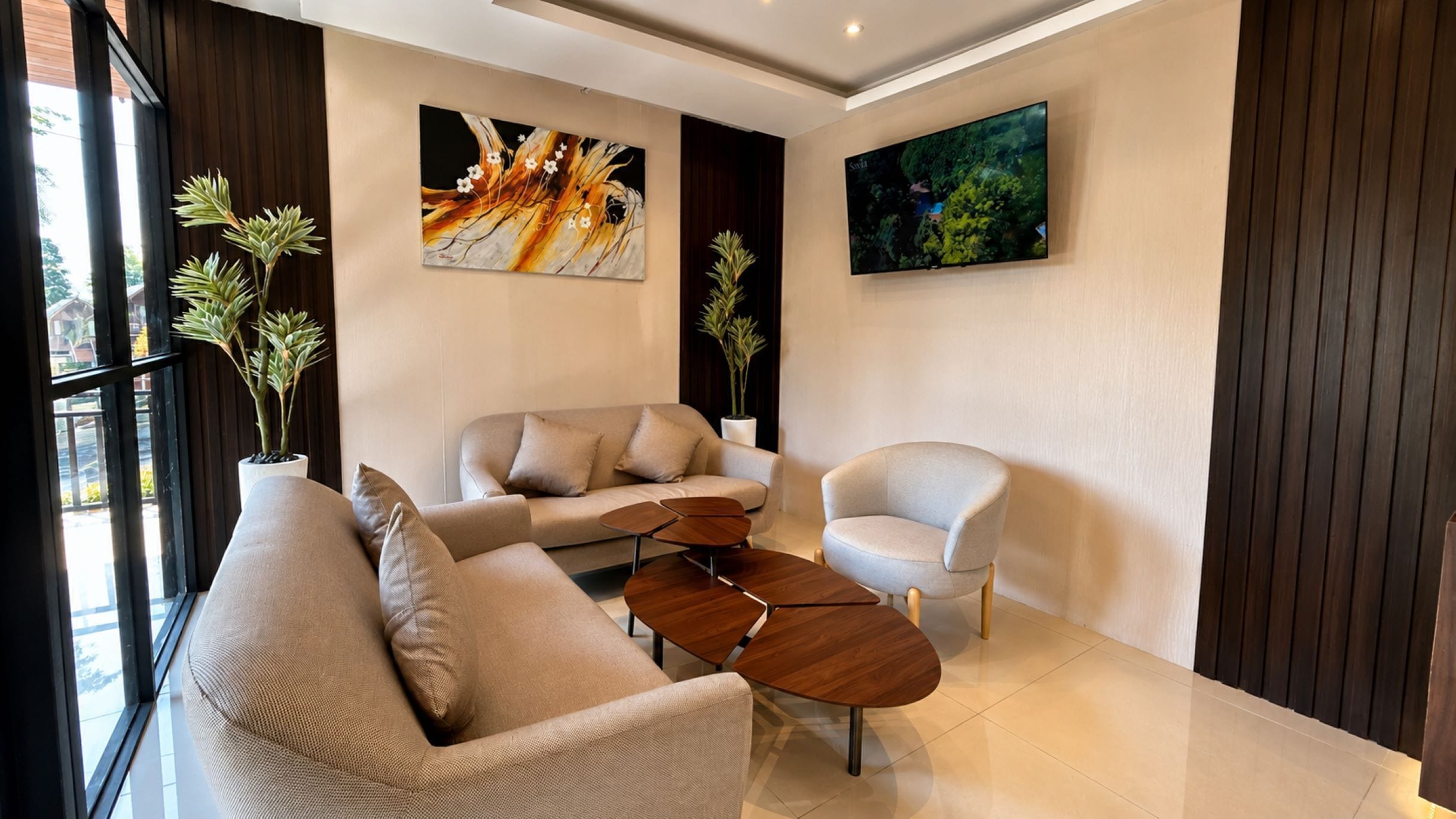 A modern living room with a sofa, armchair, and coffee table. There are two potted plants and two wall-mounted artworks.
