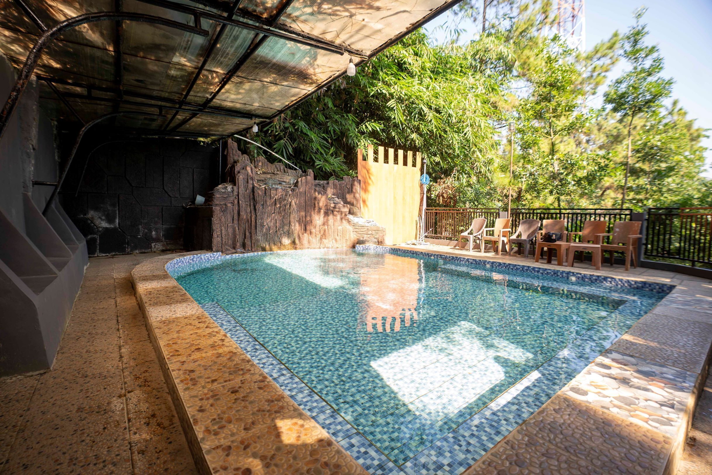 Una piscina con borde de azulejos, rodeada por una zona techada con techo de metal, un muro de piedra y una valla de madera.