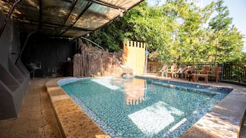 Una piscina con borde de azulejos, rodeada por una zona techada con techo de metal, un muro de piedra y una valla de madera.