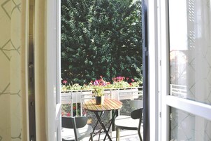 Una porta aperta che dà su un balcone con tavolo e sedie, immerso nel verde e fiorito.