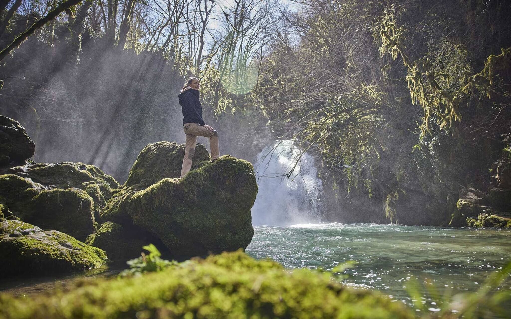 Iemand staat op een met mos begroeide rots naast een waterval in een bebost gebied.