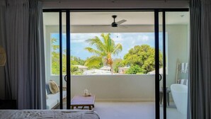 A room with a bed, a small table with a bottle and a cup, a ceiling fan, and a sliding glass door leading to a balcony with a palm tree and a view of houses. - Walk to Beach New Elegant 3BR with Pool  (Lower Carlton)