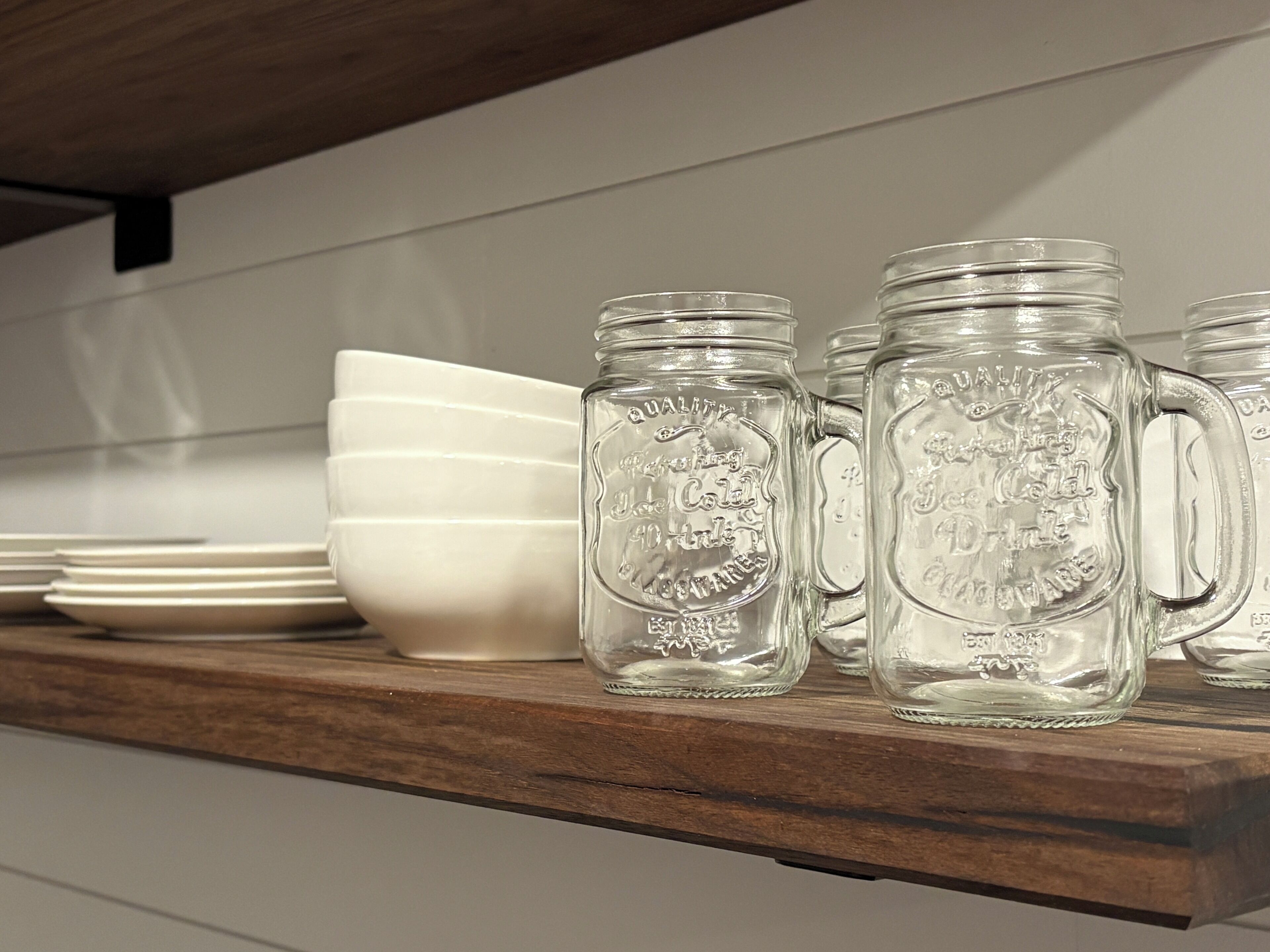 A wooden shelf with clear glass mugs and white plates.