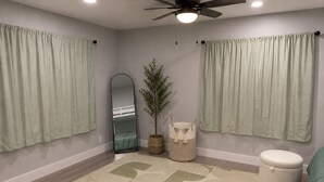 Una habitación con ventilador de techo, un espejo grande, una planta en maceta, una cesta tejida y un otoman redondo.