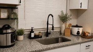 Una cocina con encimera de granito, electrodomésticos de acero inoxidable, armarios blancos y una ventana con persianas.