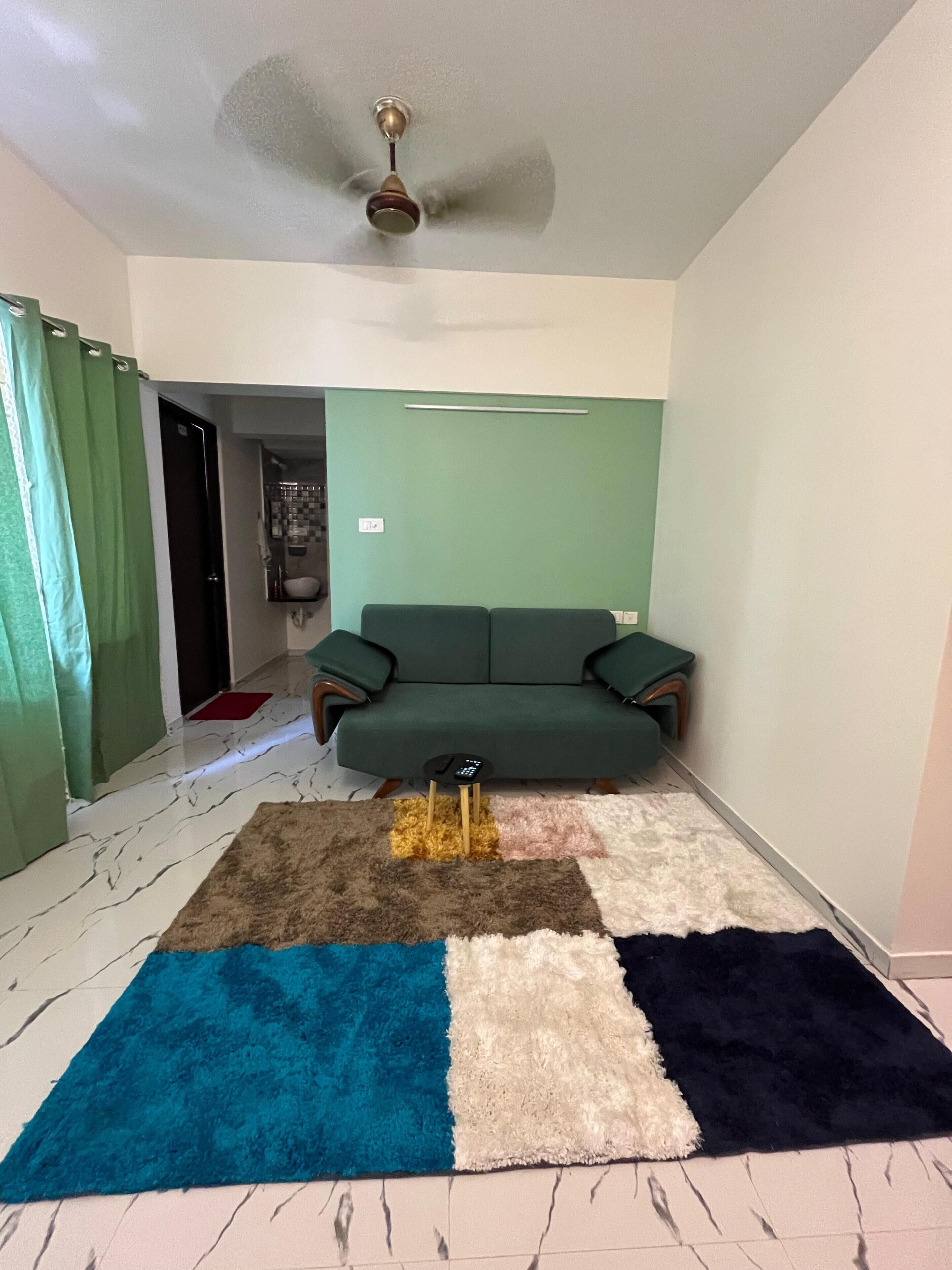 Una habitación con un sofá verde, un ventilador de techo, un tapete estampado y una cortina verde.