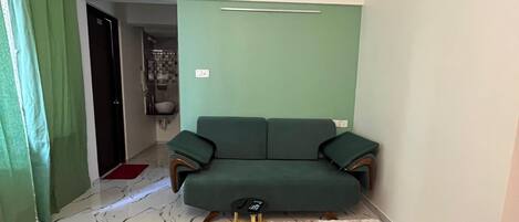 Una habitación con un sofá verde, un ventilador de techo, un tapete estampado y una cortina verde.
