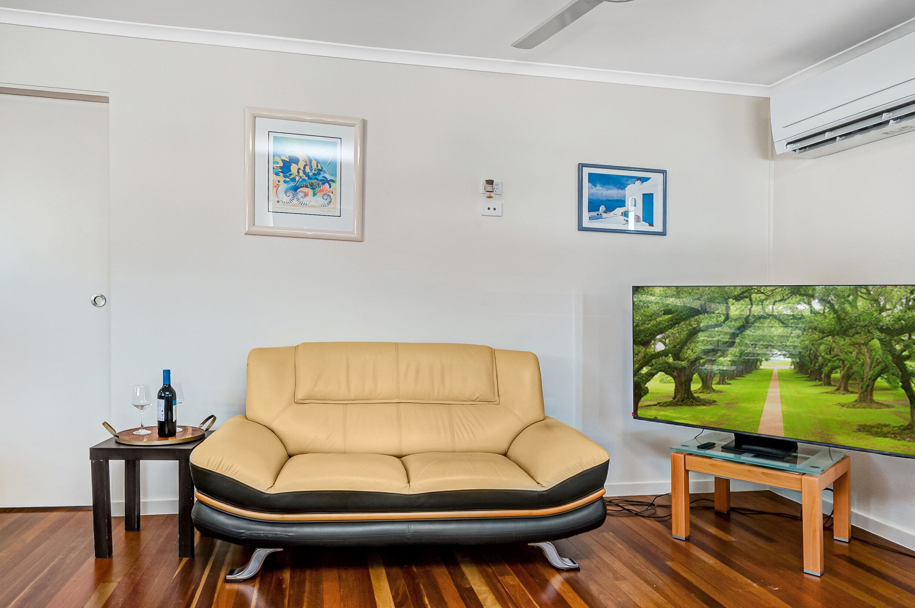 A living room with a beige sofa, a small table with a wine bottle and glass, a TV on a stand, and two framed pictures on the wall.