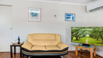 A living room with a beige sofa, a small table with a wine bottle and glass, a TV on a stand, and two framed pictures on the wall.