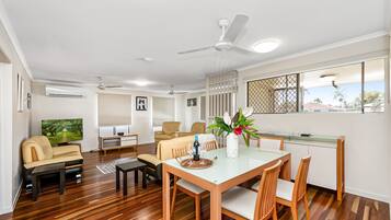 A modern living room with a sofa, TV, dining table, chairs, and a kitchen area with a window.