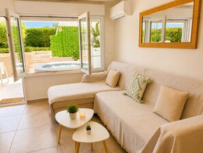 A modern living room with a sofa, a small table, and a window with a view of a garden and a hot tub. - Beachfront Jacuzzi Retreat Seaview Walk Everywhere (Saronida)