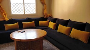 Een woonkamer met een donkere bank, gele kussens, een ronde houten salontafel en een raam met gele gordijnen.