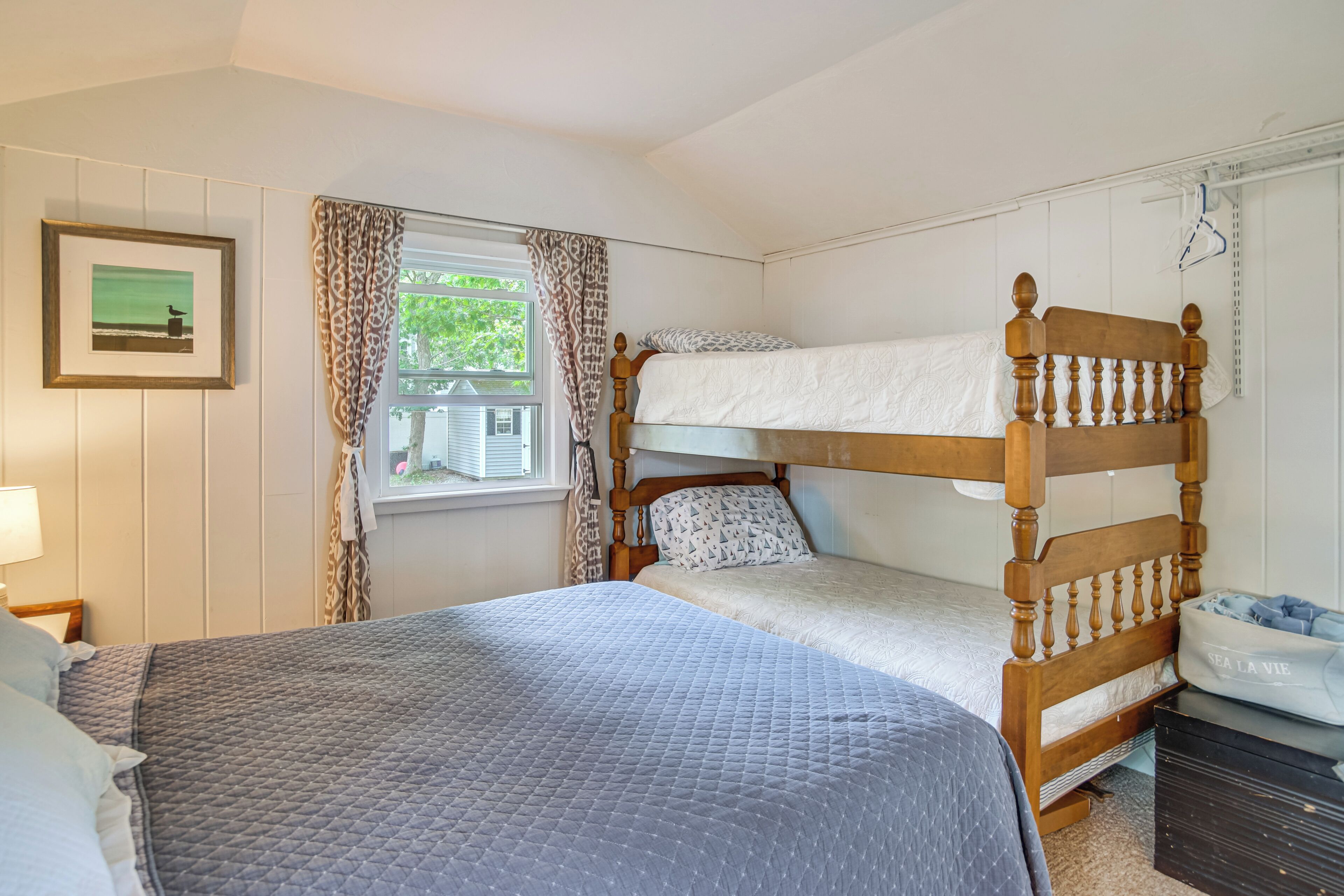 A room with a bunk bed, a single bed, a framed picture, a window with curtains, and a dresser.
