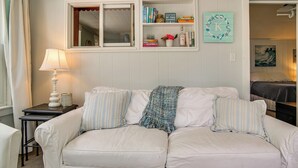 Un sofá blanco con almohadones a rayas, una mesita con una lámpara, una estantería con libros y objetos decorativos, y una letra 'K' enmarcada en la pared.