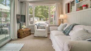 Una habitación con un sofá blanco, un cofre de madera, una silla, un televisor y una ventana con cortinas.