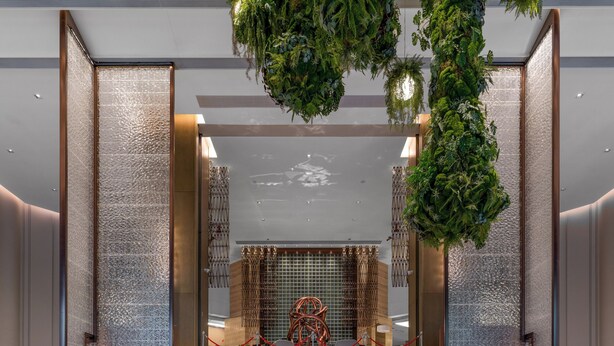 Uma escadaria imponente leva a um hall com design moderno, apresentando uma parede texturizada e plantas suspensas.