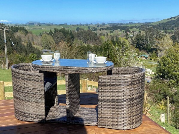 A round wicker table with a glass top and two chairs, set on a wooden deck with a scenic view of hills and trees. - Czars Rest Tiny Home (Oruanui)