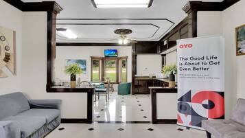A hotel lobby with a reception desk, a television, a dining area with chairs and a table, and a large OYO advertisement.