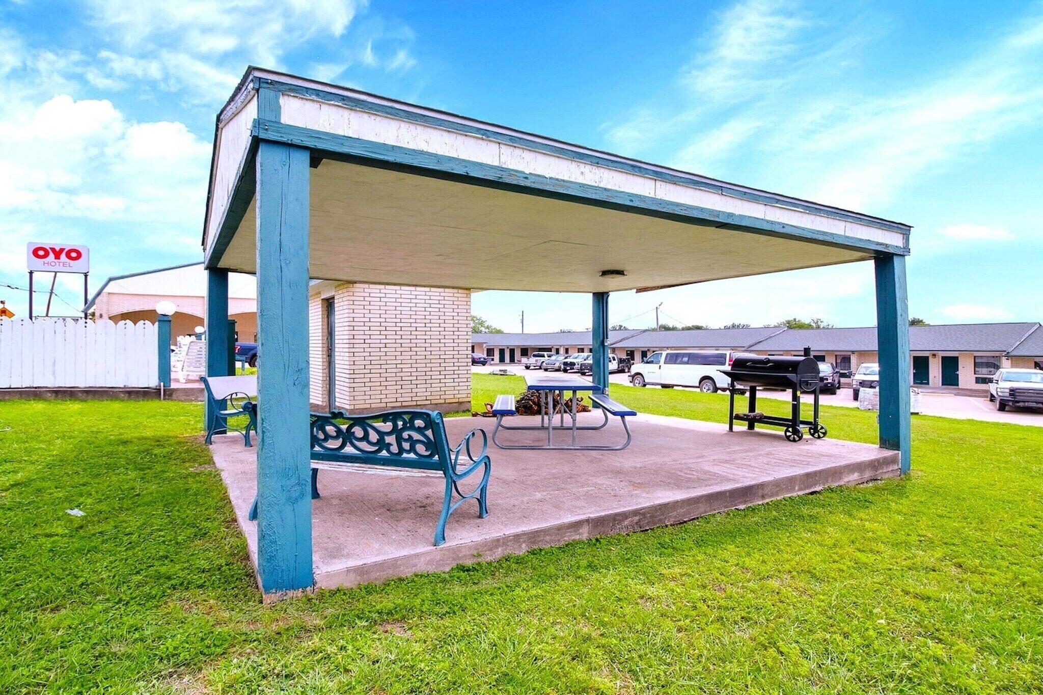 Un área de estar al aire libre cubierta con mesa de picnic y bancos, un edificio de ladrillo y un estacionamiento con coches.