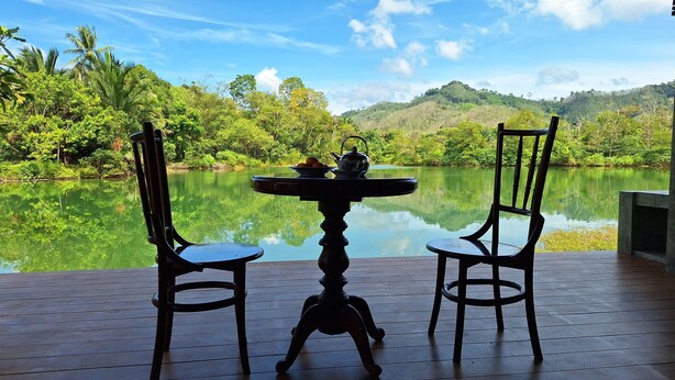 Una terraza de madera con dos sillas y una mesa, con vistas a un lago tranquilo, vegetación exuberante y colinas al fondo.