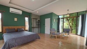 Un dormitorio con cama, escritorio, dos sillas, ventana con vista a árboles y un equipo de aire acondicionado.