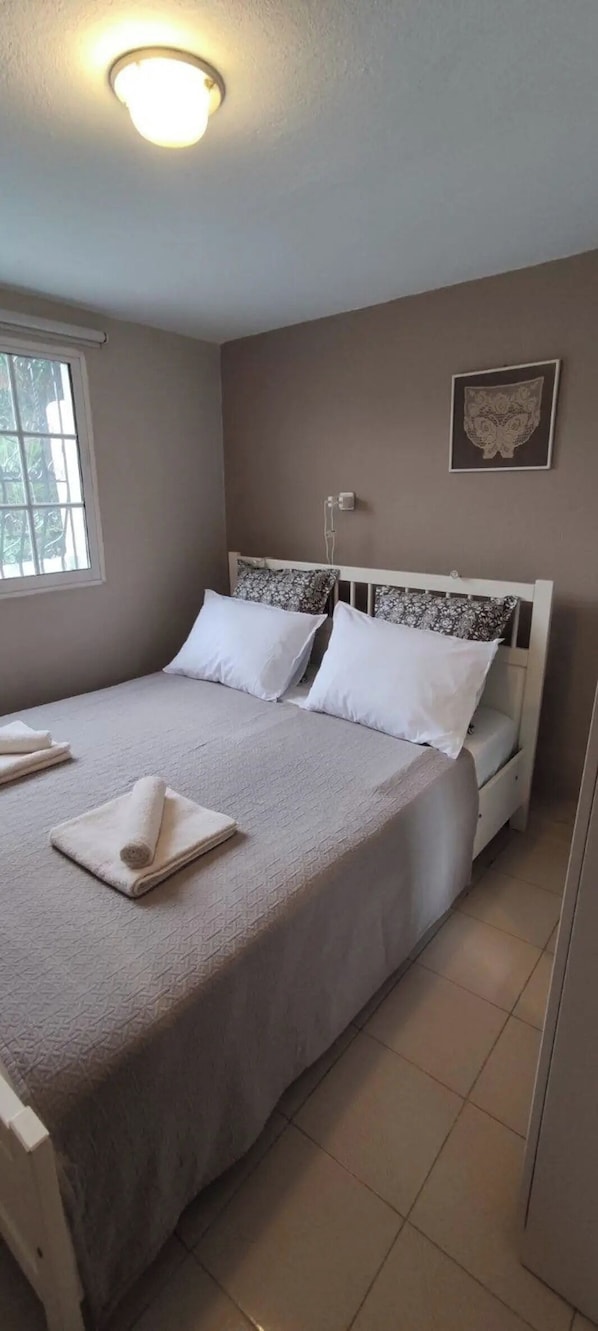 A bedroom with a bed, pillows, a bedside table, a wall-mounted light, and a window.