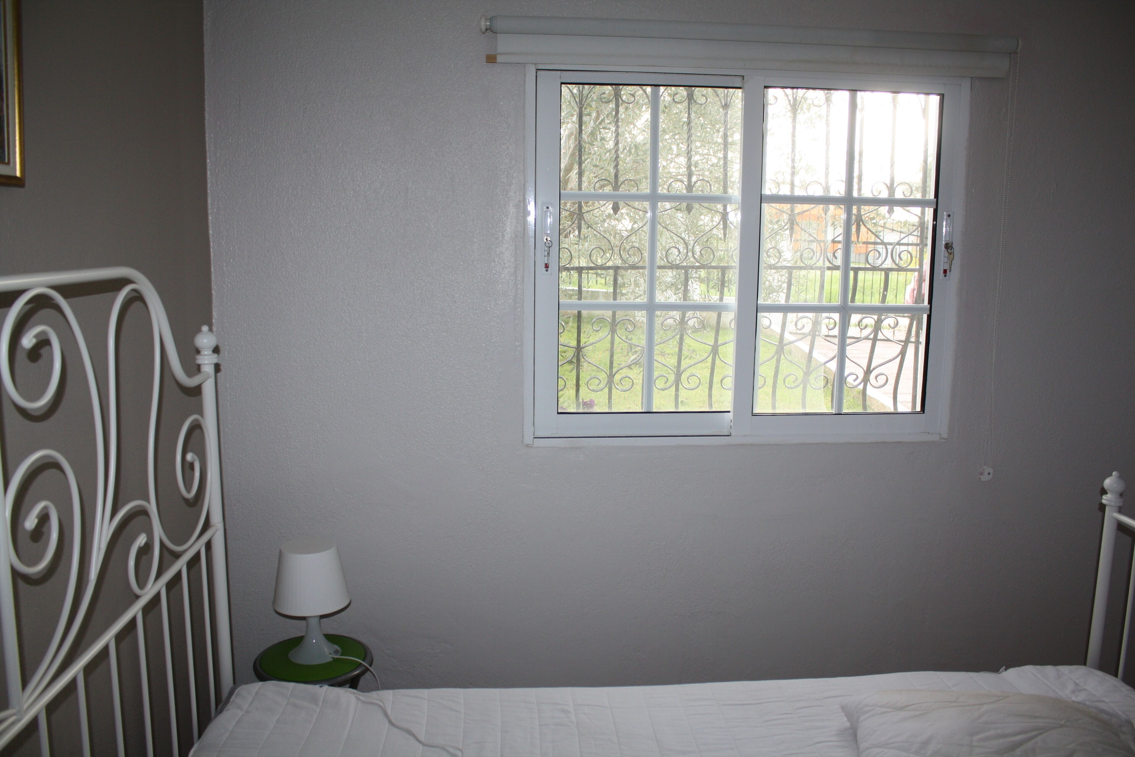 Un dormitorio con una cama individual, una ventana que da a un jardín y una mesita de noche con una lámpara.