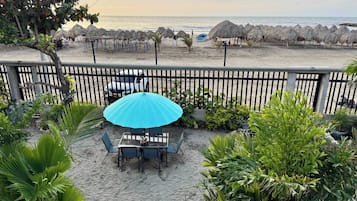 Une terrasse avec une table et des chaises, un parasol bleu et une vue sur la plage, où l’on aperçoit d’autres parasols en feuilles de palmier.