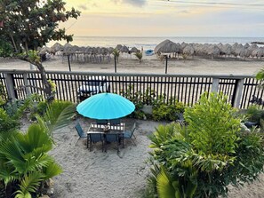 Un patio con una mesa y sillas, una sombrilla azul y vistas a la playa con sombrillas de paja.