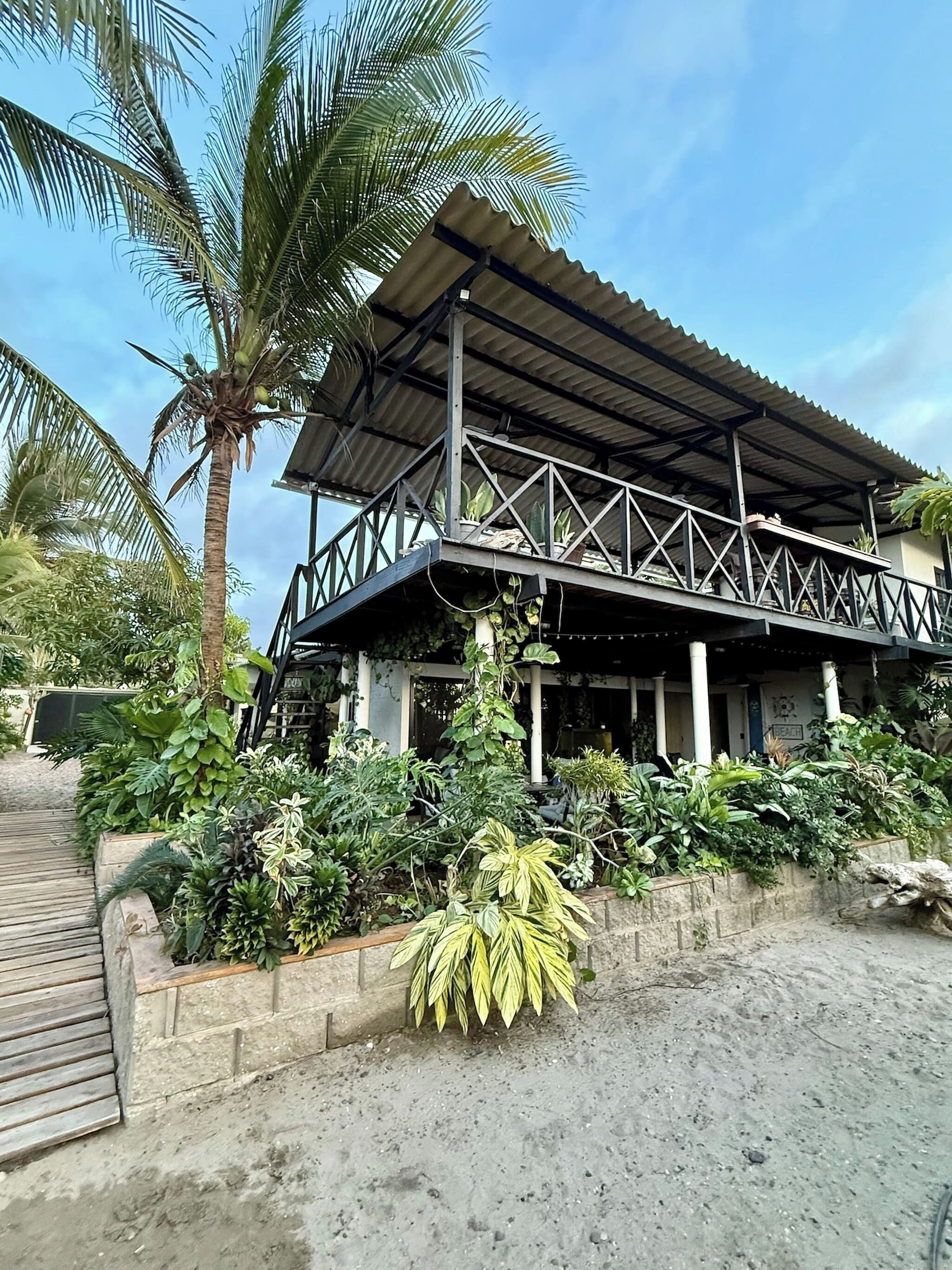 Un bâtiment à deux étages, doté d’un toit en métal et d’un balcon, entouré d’une végétation tropicale luxuriante.