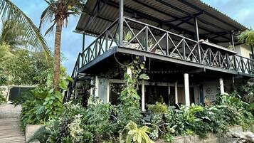 Un bâtiment à deux étages, doté d’un toit en métal et d’un balcon, entouré d’une végétation tropicale luxuriante.