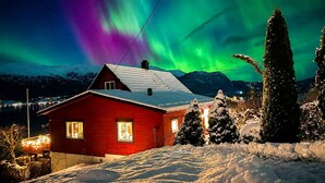Une maison rouge, dont le toit est recouvert de neige, entourée de neige et d’arbres, sous un ciel nocturne illuminé par des aurores boréales.