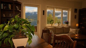 Une pièce chaleureuse avec une table en bois, des chaises, un canapé et une bibliothèque. De grandes fenêtres offrent une vue sur les montagnes et un lac.